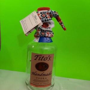 May include: A clear glass bottle of Tito's Handmade Vodka with a red and white knitted bottle cover. The bottle label features the brand name, "Tito's Handmade Vodka", and the text "Award Winning American Vodka Crafted in an Old Fashioned Pot Still" in pink lettering. The bottle also has a blue label with the text "Texas USA Bottled" in white lettering.