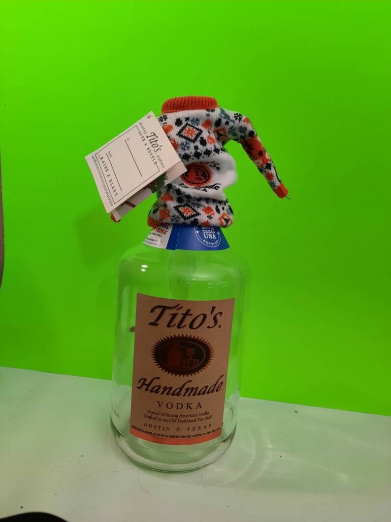 May include: A clear glass bottle of Tito's Handmade Vodka with a red and white knitted bottle cover. The bottle label features the brand name, "Tito's Handmade Vodka", and the text "Award Winning American Vodka Crafted in an Old Fashioned Pot Still" in pink lettering. The bottle also has a blue label with the text "Texas USA Bottled" in white lettering.