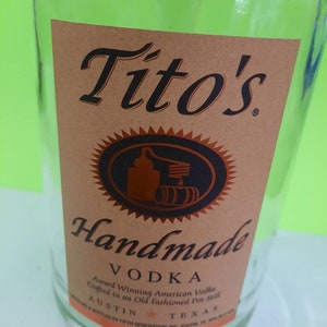 May include: A clear glass bottle with a brown label that reads "Tito's Handmade Vodka". The label also includes the text "Award Winning American Vodka Crafted in an Old Fashioned Pot Still" and "Austin, Texas".