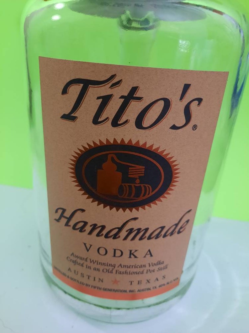 May include: A clear glass bottle with a brown label that reads "Tito's Handmade Vodka". The label also includes the text "Award Winning American Vodka Crafted in an Old Fashioned Pot Still" and "Austin, Texas".