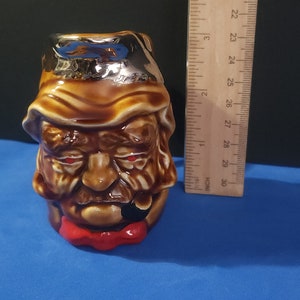 May include: A ceramic mug shaped like a man's head with a brown beard and mustache. The mug has a red bow tie and a silver rim. The man's eyes are red and he is wearing a brown hat.