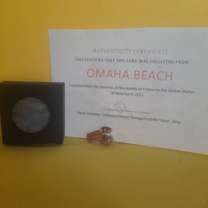 May include: A black square box with a circular design, a small glass vial with sand, and a certificate. The certificate reads "Authenticity Certificate" and states the sand was collected from Omaha Beach.