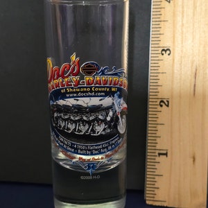 May include: A clear glass shot glass with a Harley-Davidson logo and a motorcycle graphic. The text on the glass reads "Doc's Harley-Davidson of Shawano County, WI www.docshd.com Fun Facts - 4 1950's Flathead 45ci. Engines - Black Start At Once - Built by "Doc" Aug. 08 in 1 Day? Way at Doc's to  ©2009 H-D"