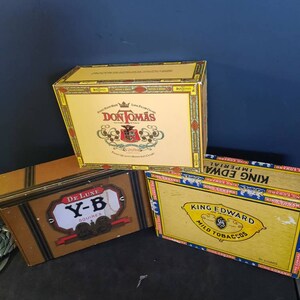 May include: Three vintage cigar boxes. The top box is yellow with black and red lettering and a crest. It says "Don Tomas" and "Fine Hand Made Long Filler Cigars". The middle box is brown with black and gold lettering and says "De Luxe Y-B Squires". The bottom box is yellow with blue and gold lettering and says "King Edward Imperial Mild Tobaccos" and "50 Cigars".