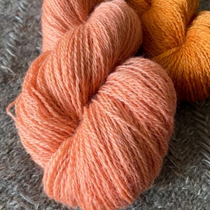 May include: Close-up of several skeins of yarn in shades of peach and orange. The yarn is soft and fluffy, with a textured appearance. The yarn is arranged on a grey, textured surface.