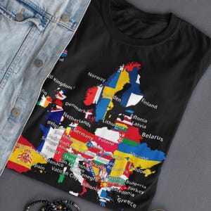 Peut inclure: Un t-shirt noir présentant une carte de l'Europe où chaque pays est rempli de son drapeau national respectif et étiqueté avec son nom en texte blanc. Le t-shirt est présenté avec une veste en jean bleu clair, un bracelet de perles et des lunettes.