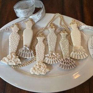 May include: Six intricately decorated, dress-shaped biscuits in shades of white and ivory, presented on a white plate. A pearl necklace and a ribbon with the word "Bride" are also visible. The biscuits feature detailed designs, including ruffles and floral patterns.