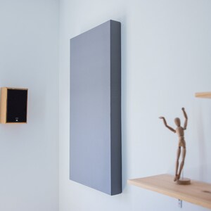 May include: Acoustic panel and speaker mounted on a white wall. The large, rectangular panel is gray, while the speaker has a wooden frame and a black front. A wooden artist's mannequin and a shelf with books are also visible.