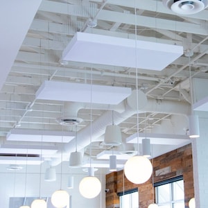 May include: A white ceiling with white rectangular acoustic panels and a white ceiling fan. A large chandelier with multiple clear glass globes hangs from the ceiling. The globes are illuminated with warm white light. The chandelier is centered in the image. The background is a white wall with a brick wall behind it.