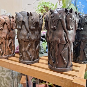 May include: Four decorative cylindrical vases, each featuring a relief of multiple horse heads and bodies. The vases are in shades of brown and black, with a textured finish. They are displayed on a wooden shelf.