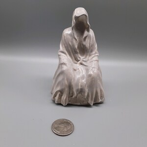 May include: A white ceramic sculpture of a hooded figure sitting with their head down. The sculpture is about 6 inches tall.