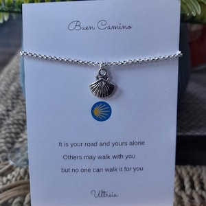 May include: A silver chain bracelet with a scallop shell pendant. The pendant hangs from a delicate chain. The card behind the bracelet says "Buen Camino" and includes a quote about the journey.