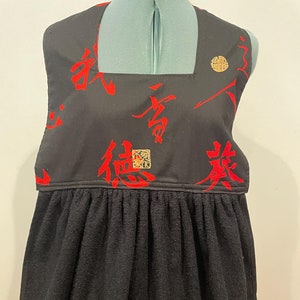 以下が含まれることがあります： 赤い中国語の文字と金のアクセントが付いた黒いキッチンタオル。タオルはギャザーの入った裾とスクエアネックラインです。