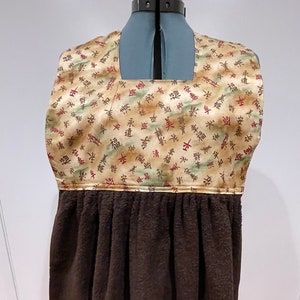 以下が含まれることがあります： 正方形の首と茶色のテリークロス地の底を持つ茶色とベージュのキッチンタオル。上部は、茶色、緑、赤の色合いの日本の文字とスタイリッシュな木の模様で飾られています。