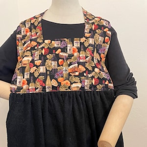 May include: A black apron with a towel bottom. The top portion features a colorful pattern of wine bottles, grapes, and cheese. The apron is worn over a black long-sleeved shirt.