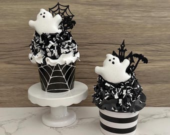 May include: Two Halloween-themed decorative cupcakes. One cupcake sits on a white pedestal and features a black and white spiderweb design. The other cupcake has black and white stripes. Both have white ghost toppers and black decorative accents.