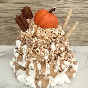 May include: A decorative faux dessert featuring a pumpkin-shaped element, cinnamon sticks, and two straws. The dessert has layers of white and brown frosting, topped with nuts. The overall color palette includes shades of brown, white, and orange.