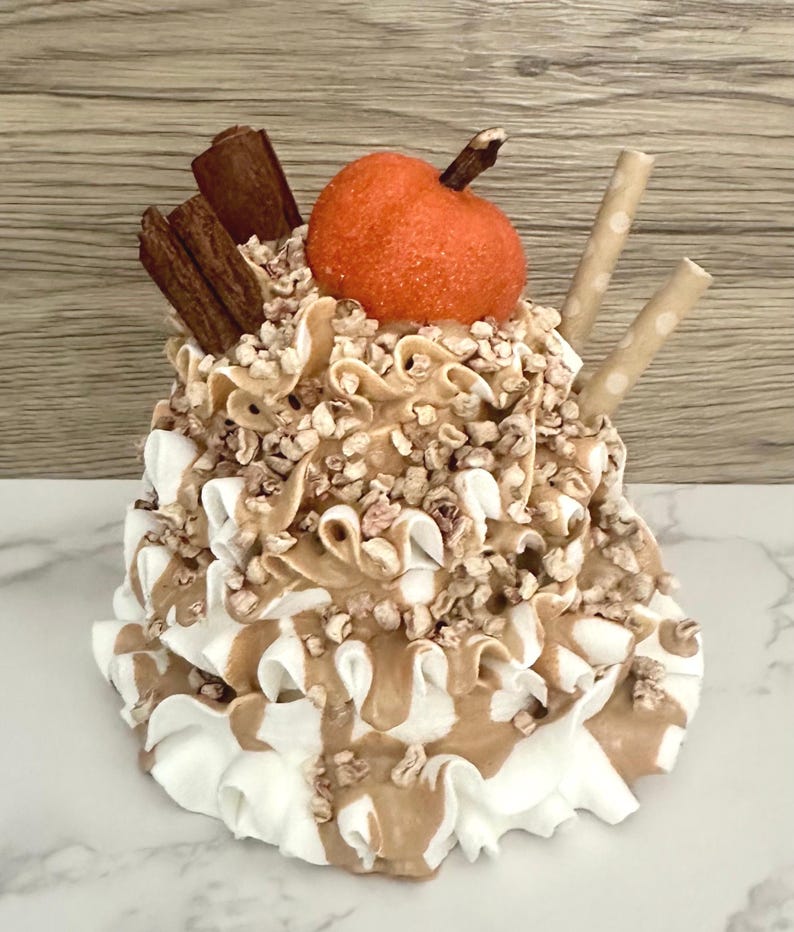 May include: A decorative faux dessert featuring a pumpkin-shaped element, cinnamon sticks, and two straws. The dessert has layers of white and brown frosting, topped with nuts. The overall color palette includes shades of brown, white, and orange.