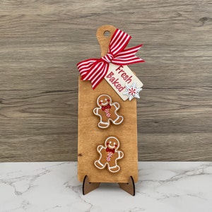 May include: Wooden cutting board-shaped sign with two gingerbread men, a red and white striped bow, and a tag that reads "Fresh Baked." A white snowflake is attached to the tag. The sign is propped up on a wooden stand.