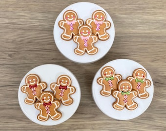 May include: Three white ceramic containers each topped with gingerbread man cookies. The cookies have smiling faces and are decorated with pink, red, and green icing. The cookies are arranged in groups of three.