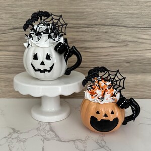 Puede incluir: Dos tazas de cer&aacute;mica con forma de calabaza con asas negras y caras de jack-o'-lantern. Una es blanca y la otra naranja. Ambas est&aacute;n cubiertas con crema batida, chispas y decoraciones de Halloween, incluyendo murci&eacute;lagos y telara&ntilde;as. Una taza est&aacute; sobre un pedestal blanco.