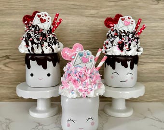 May include: Three marshmallow-shaped decorative items with smiling faces, topped with whipped cream, chocolate drizzle, and Valentine's Day embellishments. Each sits on a white pedestal. Red and pink accents.