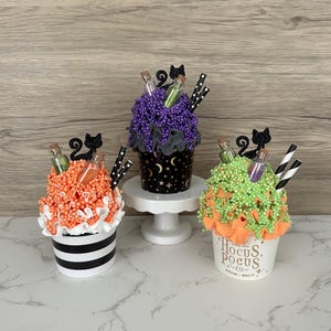 May include: Three decorative Halloween-themed cups with colorful foam toppings. Each cup features a different color of foam, decorative bottles, black cat cutouts, and striped straws. One cup has the text "Hocus Pocus Co."