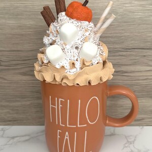 May include: A decorative fall-themed mug filled with whipped cream, marshmallows, and a small orange pumpkin. The mug is a warm orange color and has the words "HELLO FALL" in white lettering. Cinnamon sticks and striped straws add to the festive look.