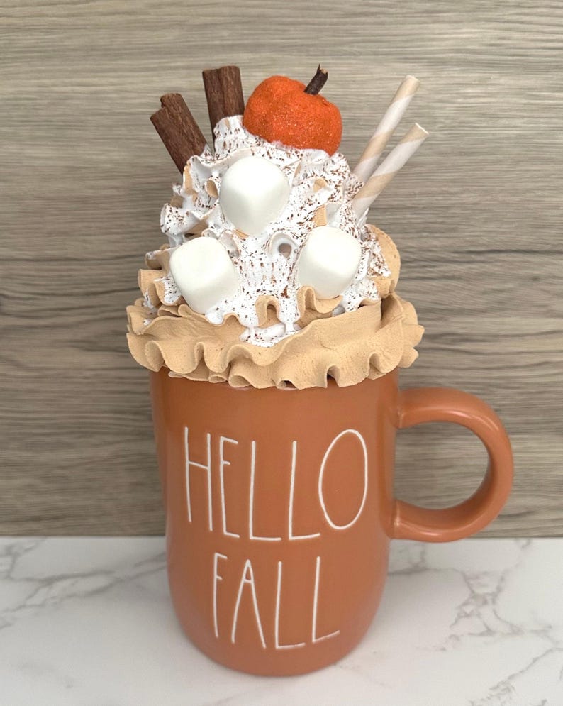 May include: A decorative fall-themed mug filled with whipped cream, marshmallows, and a small orange pumpkin. The mug is a warm orange color and has the words "HELLO FALL" in white lettering. Cinnamon sticks and striped straws add to the festive look.