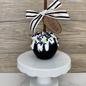 Puede incluir: Una manzana de caramelo negra cubierta de purpurina, con glaseado blanco y chispas, exhibida sobre un pedestal blanco. Un palito de madera est&aacute; coronado con un lazo a rayas blanco y negro y una etiqueta de madera que dice "poison".