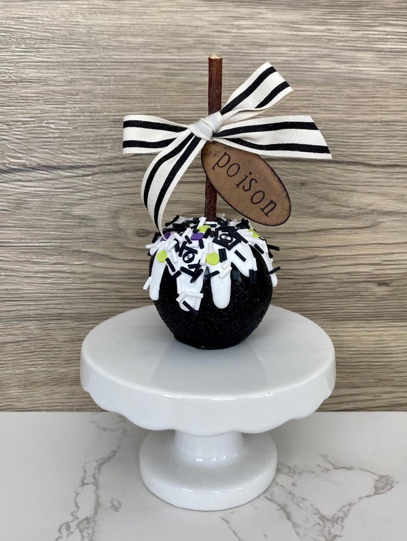 Puede incluir: Una manzana de caramelo negra cubierta de purpurina, con glaseado blanco y chispas, exhibida sobre un pedestal blanco. Un palito de madera est&aacute; coronado con un lazo a rayas blanco y negro y una etiqueta de madera que dice "poison".