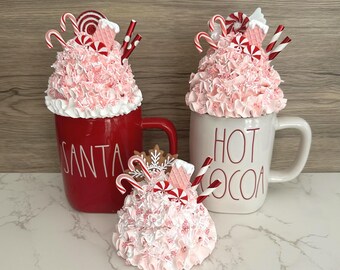 May include: Three decorative mugs with festive holiday themes. One red mug says "SANTA" and another white mug says "HOT COCOA". Each mug is topped with pink, ruffled frosting, candy canes, and other holiday-themed decorations. A smaller decoration sits in front.