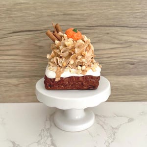 May include: A square-shaped dessert topped with tan frosting, chopped nuts, two cinnamon sticks, and a small orange pumpkin decoration. The dessert sits on a white pedestal stand with a scalloped edge, featuring a dark brown base layer.