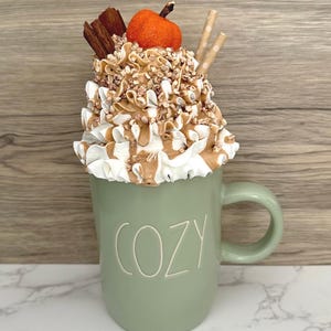 May include: A sage green mug with the word "COZY" in white lettering, topped with whipped cream, caramel drizzle, and a small orange pumpkin. Two paper straws and cinnamon sticks complete the fall-themed design.