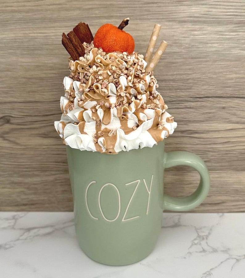 May include: A sage green mug with the word "COZY" in white lettering, topped with whipped cream, caramel drizzle, and a small orange pumpkin. Two paper straws and cinnamon sticks complete the fall-themed design.