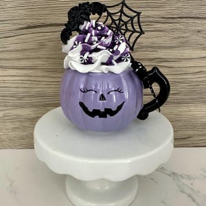 Puede incluir: Una taza morada con forma de calabaza con una cara sonriente de jack-o'-lantern, cubierta con crema batida blanca, chispas moradas y una decoraci&oacute;n negra de murci&eacute;lago y telara&ntilde;a. La taza est&aacute; sobre un pedestal blanco, ideal para Halloween.