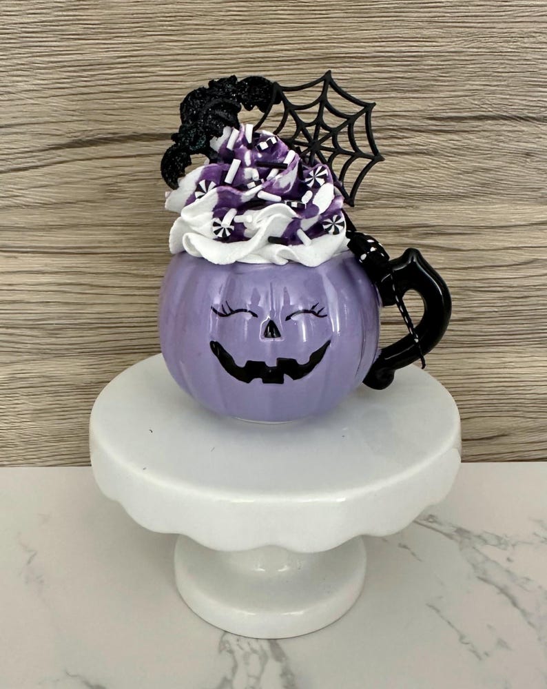 Puede incluir: Una taza morada con forma de calabaza con una cara sonriente de jack-o'-lantern, cubierta con crema batida blanca, chispas moradas y una decoraci&oacute;n negra de murci&eacute;lago y telara&ntilde;a. La taza est&aacute; sobre un pedestal blanco, ideal para Halloween.