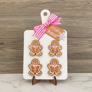 May include: A white wooden cutting board with a pink and white striped bow and a tag that says "Fresh Baked." Four gingerbread men cookies with pink bow ties are attached to the board. The board is propped up on a wooden stand.