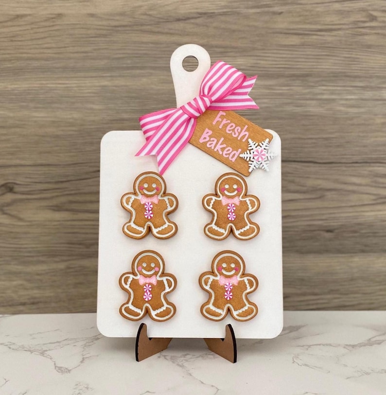May include: A white wooden cutting board with a pink and white striped bow and a tag that says "Fresh Baked." Four gingerbread men cookies with pink bow ties are attached to the board. The board is propped up on a wooden stand.
