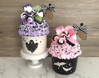 May include: Two Halloween-themed decorative cupcake-shaped containers. One is light pink with a ghost design, the other is black with bats. Both have candy and a "TREAT" tag.
