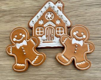 Könnte beinhalten: Lebkuchenhaus und Lebkuchenmänner. Das Haus hat weiße Glasur und silberne Perlenakzente. Die Lebkuchenmänner haben weiße Fliegen und silberne Knöpfe. Auf einer Holzoberfläche.