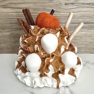 May include: A decorative dessert sculpture featuring white whipped cream, brown caramel drizzle, and three large white marshmallows. The sculpture is topped with an orange pumpkin, cinnamon sticks, and wafer sticks. The base is white with a ruffled edge.