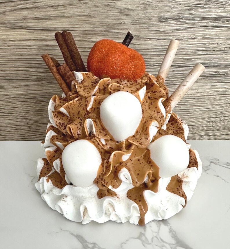 May include: A decorative dessert sculpture featuring white whipped cream, brown caramel drizzle, and three large white marshmallows. The sculpture is topped with an orange pumpkin, cinnamon sticks, and wafer sticks. The base is white with a ruffled edge.