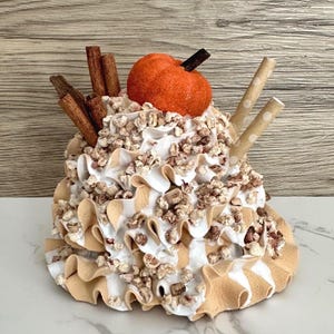 May include: A decorative fall-themed dessert with a pumpkin on top, surrounded by whipped cream, cinnamon sticks, and two paper straws. The base has a ruffled design with white and tan stripes. The dessert is decorated with small, brown, textured pieces.