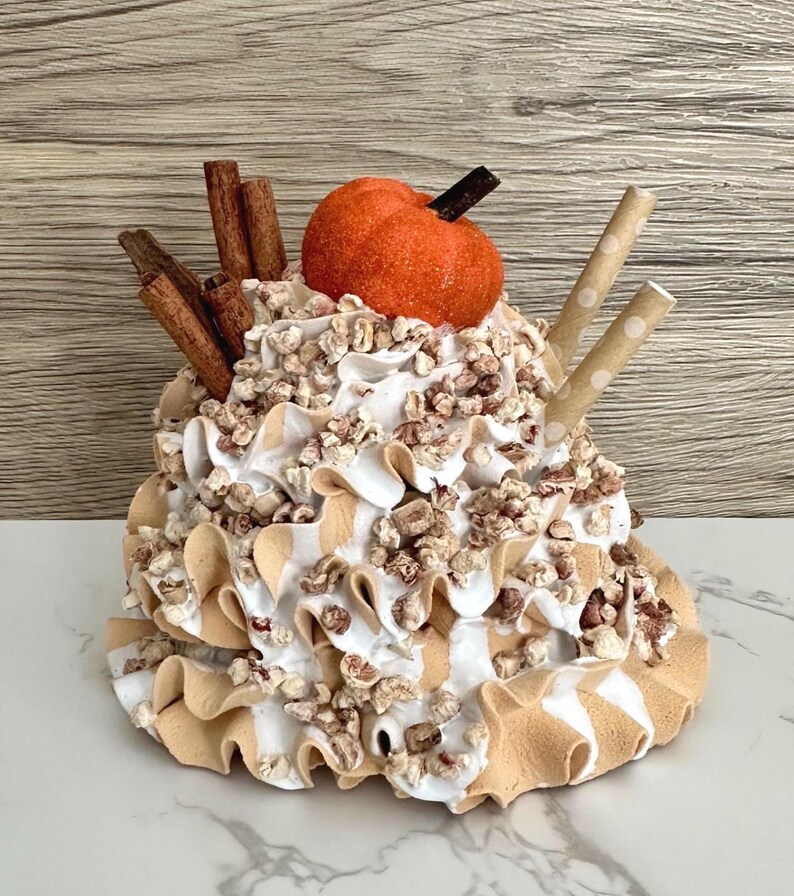 May include: A decorative fall-themed dessert with a pumpkin on top, surrounded by whipped cream, cinnamon sticks, and two paper straws. The base has a ruffled design with white and tan stripes. The dessert is decorated with small, brown, textured pieces.