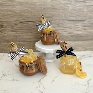 May include: Wooden honey pots and a glass jar filled with honey. Each pot has a wooden honey dipper with a bee on top and a decorative bow. The honey pots are decorated with small bee figurines.