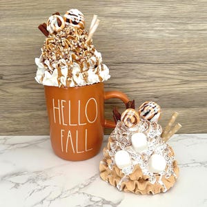 May include: An orange ceramic mug with the words "HELLO FALL" in white, filled with whipped cream, caramel drizzle, and toppings. Beside it is a smaller, similar decorative item. The items are on a white and gray surface.