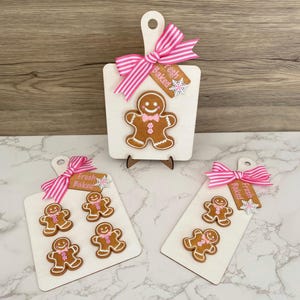 May include: Wooden cutting boards with gingerbread man designs. Each board features a pink and white striped bow and a tag that says "Fresh Baked". The gingerbread men have white icing details and pink bow ties.