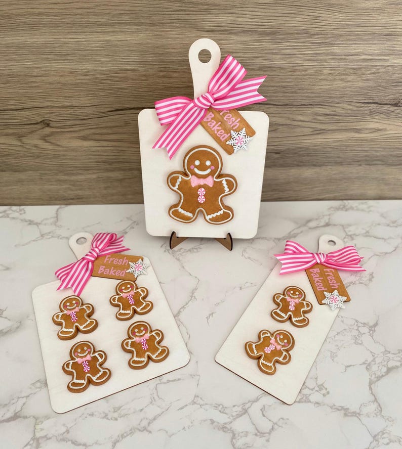 May include: Wooden cutting boards with gingerbread man designs. Each board features a pink and white striped bow and a tag that says "Fresh Baked". The gingerbread men have white icing details and pink bow ties.