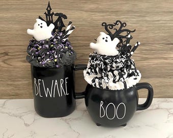 May include: Two black Halloween mugs with white text that reads "BEWARE" and "BOO". Each mug is topped with a white ghost figure, black and purple sprinkles, and decorative accents. The mugs are on a white surface.